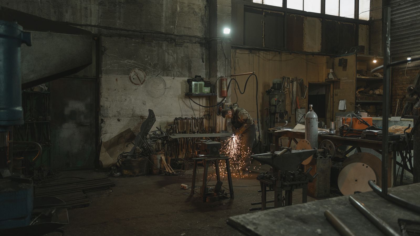Wide view of a welder at work with sparks flying in a dim workshop, face fully covered by a helmet